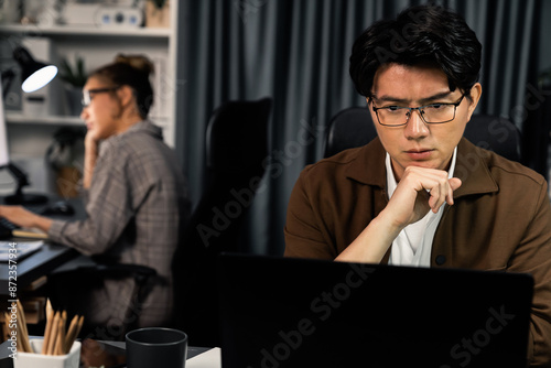 Businessman with stressful face working on laptop waiting email while coworker trading stock exchange investment at late night time at modern office, working on startup company project job. Infobahn.