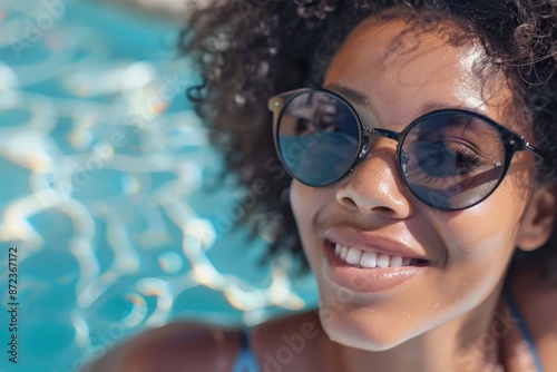 A woman with curly hair and sunglasses smiles while enjoying a sunny day by the pool, emphasizing a relaxing summertime ambiance, with sparkling blue water in the background.