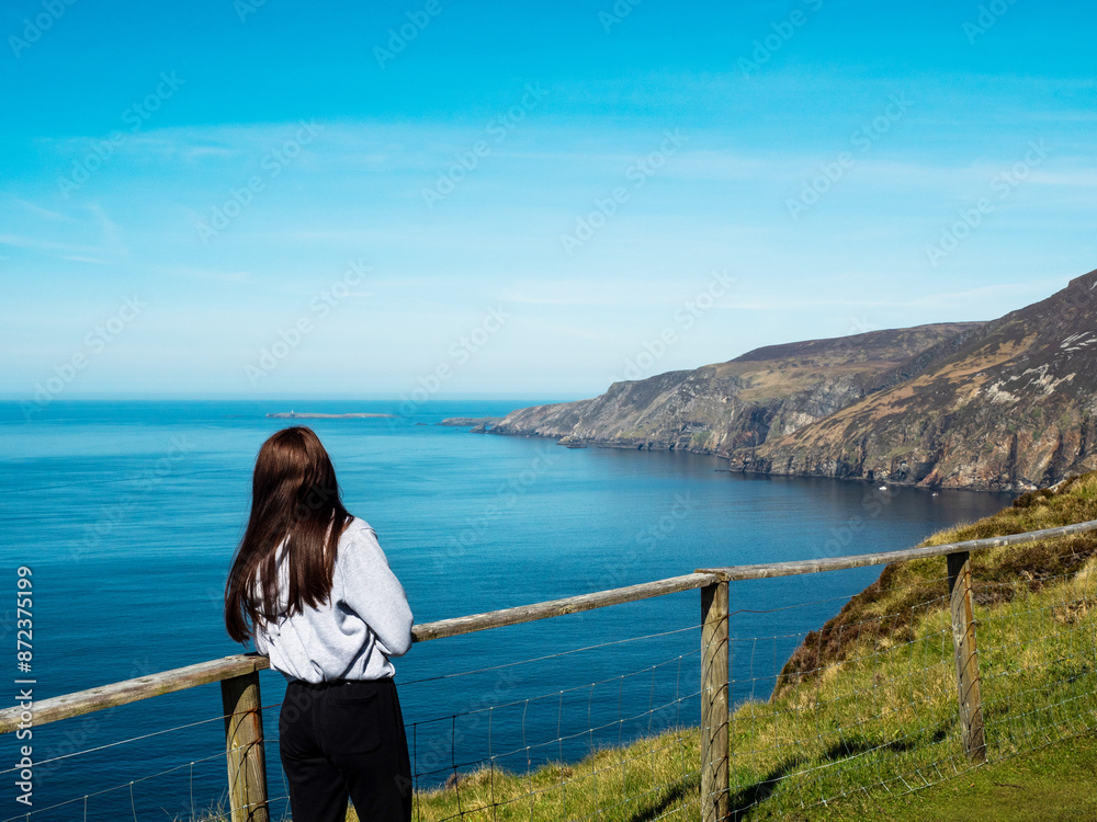 Young teenager girl exploring beautiful Sliabh Liag, county Donegal, Ireland. Travel and tourism. Warm sunny day. Stunning Irish nature landscape scene in the background. Young teenager girl exploring beautiful Sliabh Liag, county Donegal, Ireland. Travel and tourism. Warm sunny day. Stunning Irish nature landscape scene in the background.