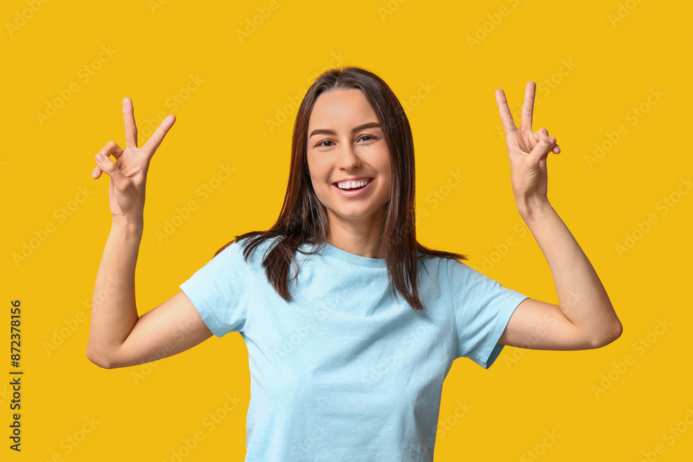 Beautiful young woman showing victory gesture on yellow background Beautiful young woman showing victory gesture on yellow background