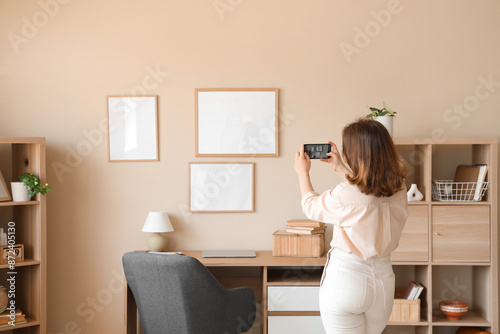 Young woman taking picture of hanging blank frames on beige wall at home