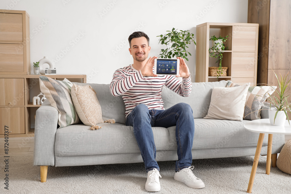 Young man with smart home security system control panel sitting on sofa in room Young man with smart home security system control panel sitting on sofa in room