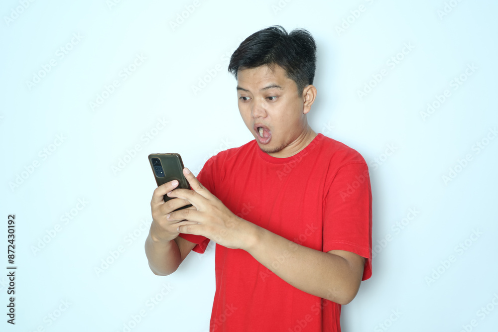 Asian man showing shocking expression looking at smartphone. Wearing red t-shirt Asian man showing shocking expression looking at smartphone. Wearing red t-shirt