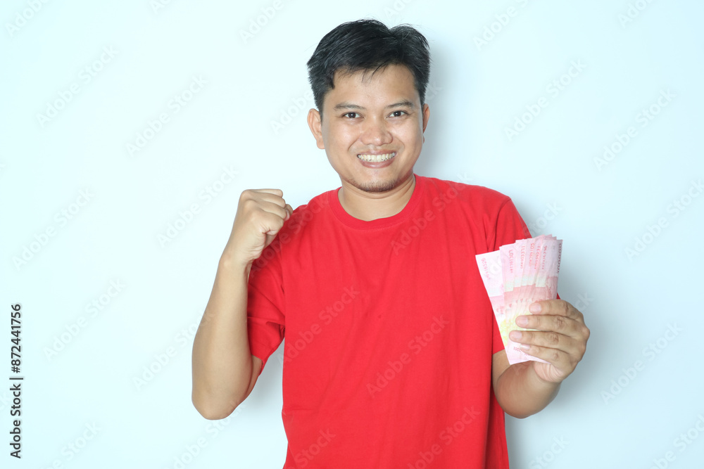 Young Asian man showing excited face expression with fist clenched while holding paper money. Wearing red t-shirt