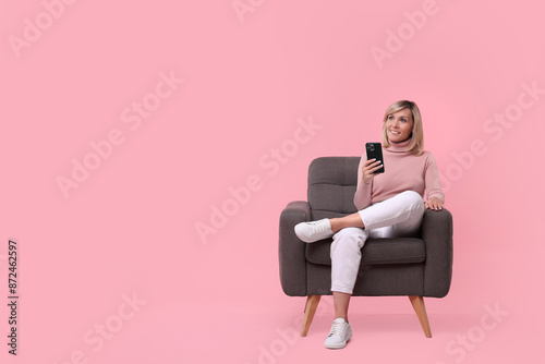 Happy woman with phone on armchair against pink background, space for text