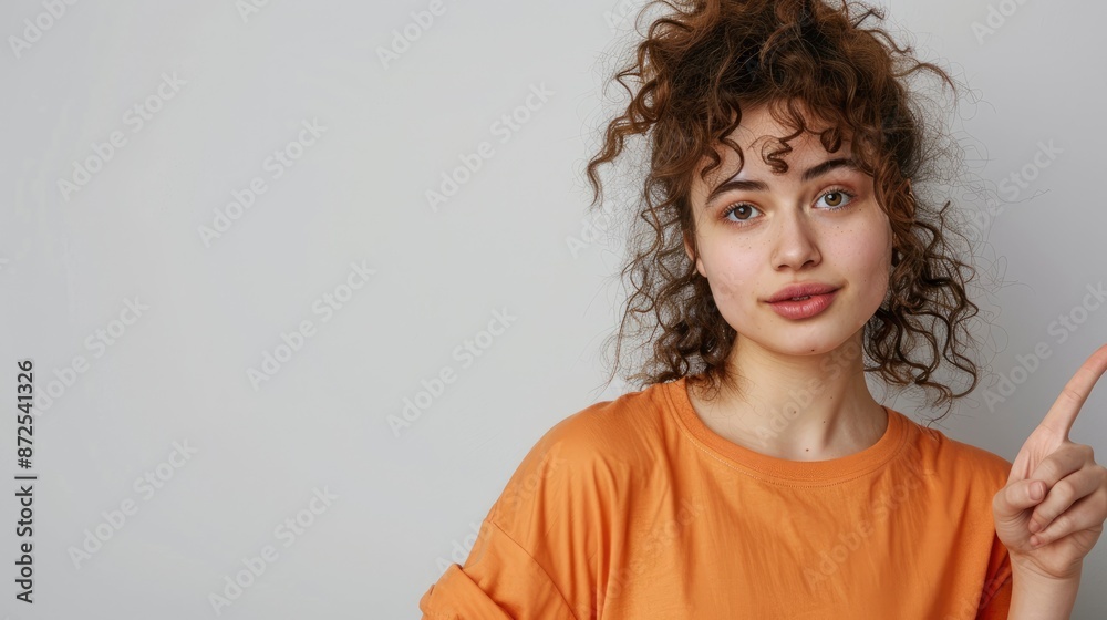 Photo Of A Young Woman With Curly Hair Pointing To The Side On A White Background, Wearing An Orange T-Shirt Photo Of A Young Woman With Curly Hair Pointing To The Side On A White Background, Wearing An Orange T-Shirt