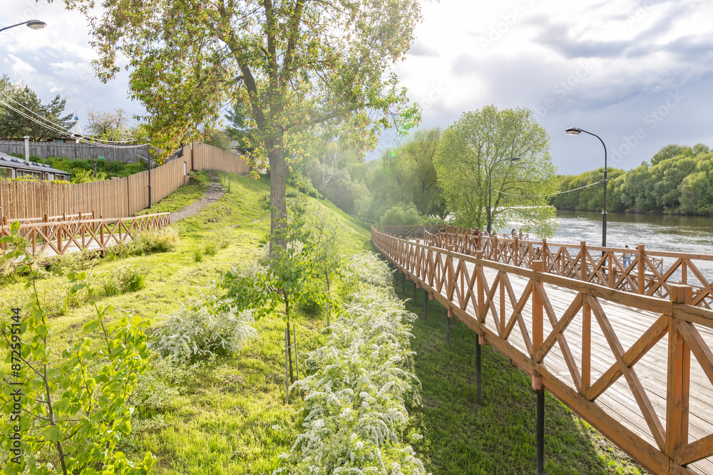Obraz premium A wooden walkway runs along the edge of a river, with green foliage and trees lining the path. Sunlight streams through the leaves, creating a dappled effect on the ground..