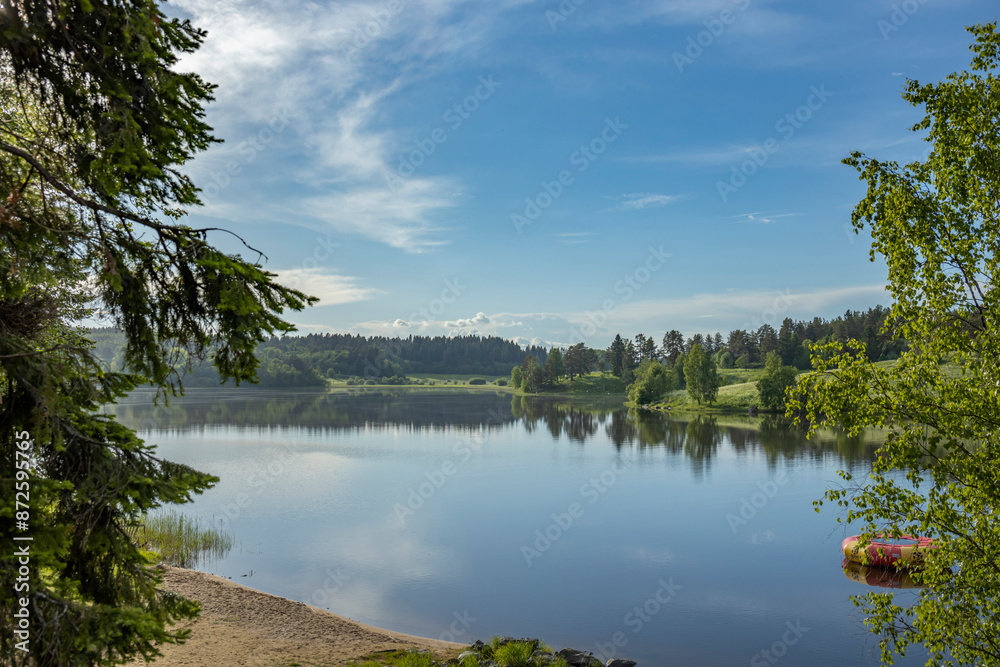 Obraz premium A still lake reflecting the blue sky and fluffy white clouds, framed by a lush green forest. The tranquility of the scene is further emphasized by the bright sunlight and calm water.