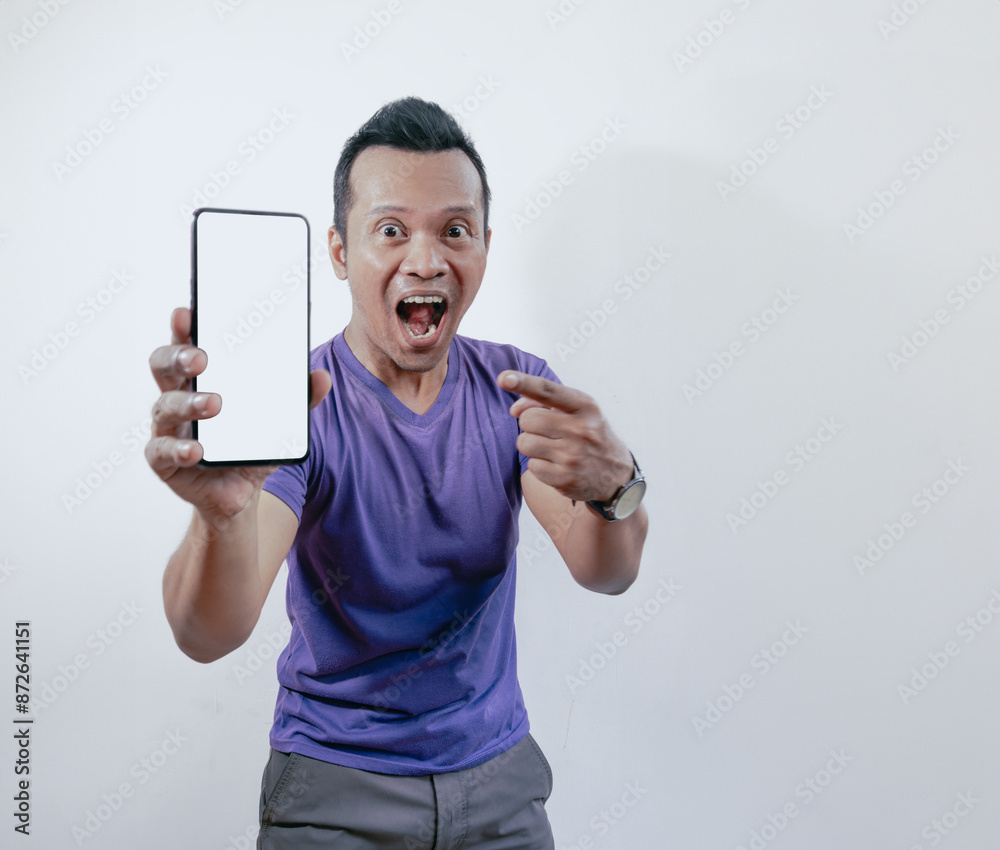 Shocked Asian man pointing and holding smartphone with blank screen, isolated on white background. Shocked Asian man pointing and holding smartphone with blank screen, isolated on white background.