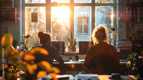 Two people working on computers in sunlit room.
