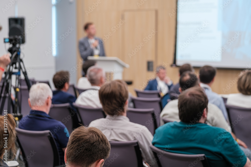 Business professionals attending a tech conference presentation with focus on the speaker and audience interaction. Business professionals attending a tech conference presentation with focus on the speaker and audience interaction.