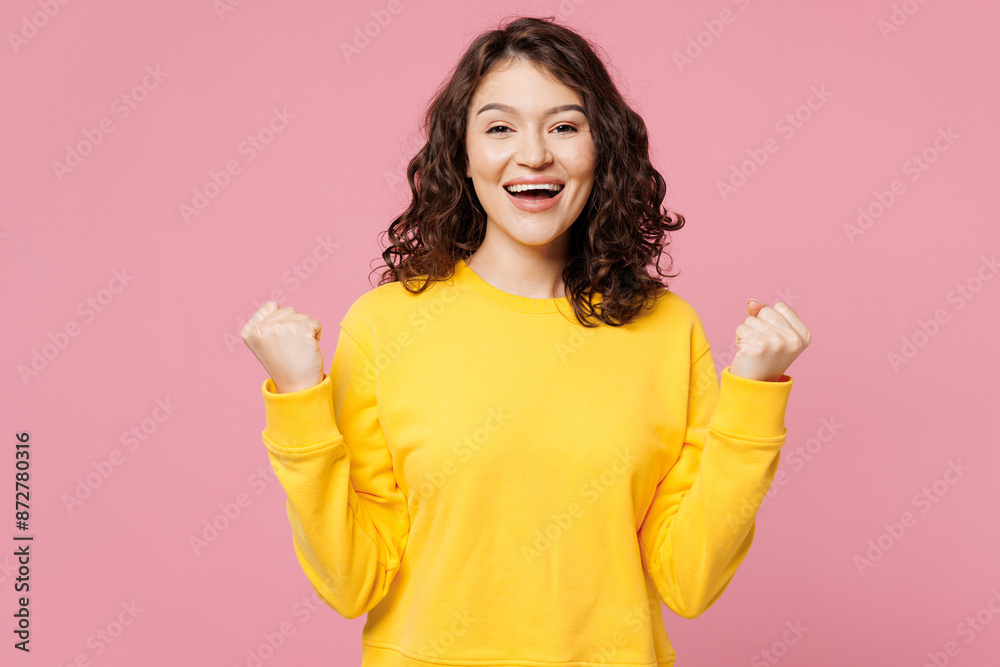 Young fun excited woman she wears yellow sweatshirt casual clothes doing winner gesture celebrate clenching fists say yes isolated on pastel light pink background studio portrait. Lifestyle concept. Young fun excited woman she wears yellow sweatshirt casual clothes doing winner gesture celebrate clenching fists say yes isolated on pastel light pink background studio portrait. Lifestyle concept.