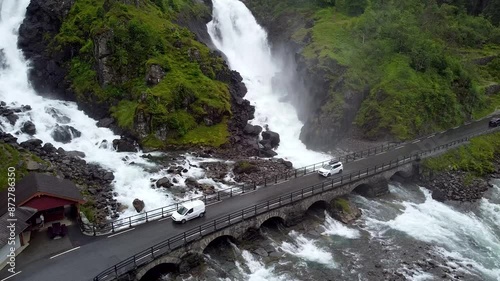 Drone footage of Låtefossen Waterfall cascading beside a stone bridge in Norway. Cars pass over the bridge, enhancing the scenic and powerful natural beauty. photo