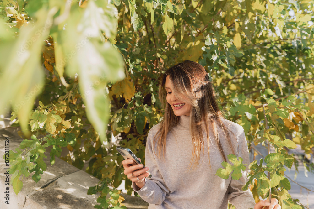A woman is sitting under a tree and looking at her cell phone. She is smiling and she is enjoying herself. A woman is sitting under a tree and looking at her cell phone. She is smiling and she is enjoying herself.
