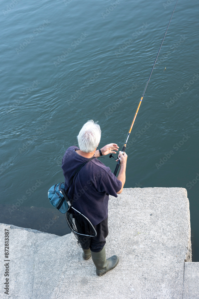 An individual with white hair is engaged in fishing on a concrete pier, overlooking a calm and serene body of water. The person is equipped with fishing gear and appears focused. An individual with white hair is engaged in fishing on a concrete pier, overlooking a calm and serene body of water. The person is equipped with fishing gear and appears focused.