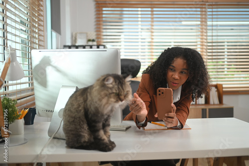 Smiling African American freelancer taking picture of her lovely cat at home office desk