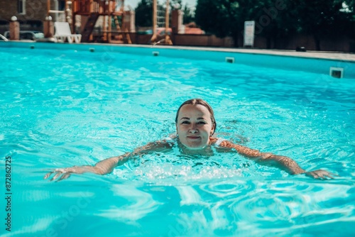 A woman is swimming in a pool. She is smiling and enjoying herself. The pool is surrounded by trees and there are chairs and a bench nearby. © svetograph