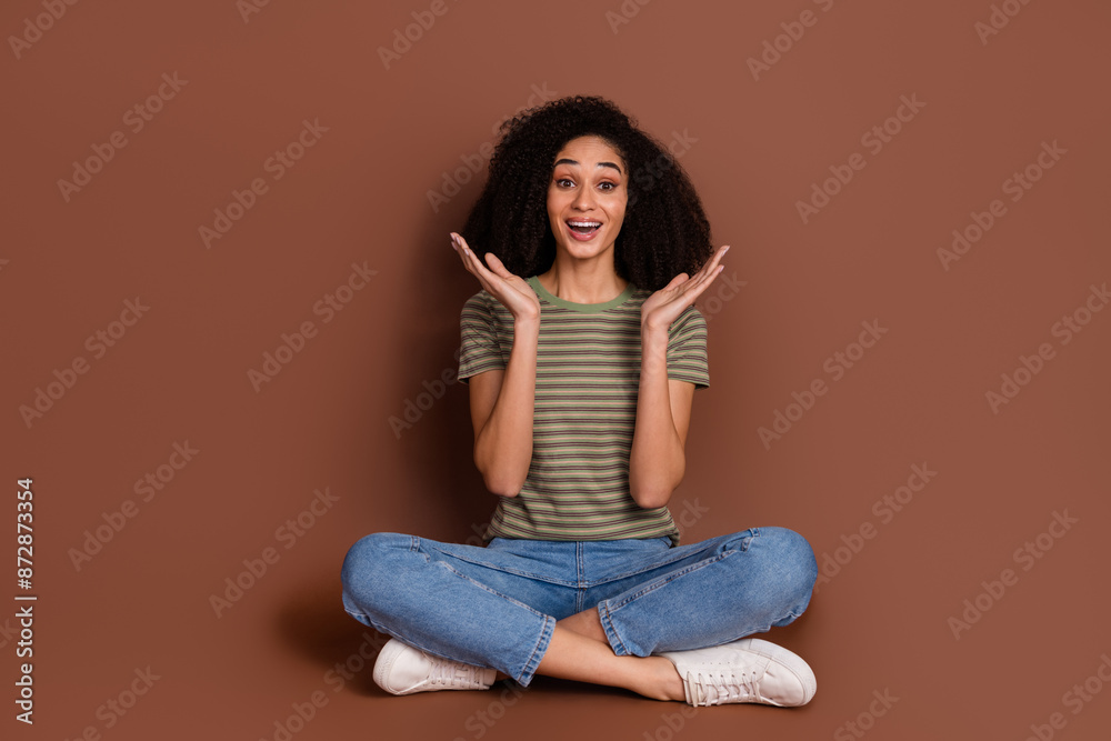 Full length photo of cheerful excited lady dressed khaki t-shirt sitting legs crossed rising arms empty space isolated brown color background Full length photo of cheerful excited lady dressed khaki t-shirt sitting legs crossed rising arms empty space isolated brown color background