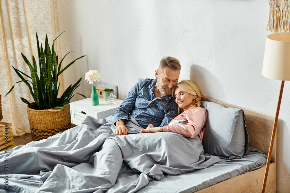 A man and woman, in cozy homewear, lovingly embrace while laying on a bed under a soft blanket in their bedroom. A man and woman, in cozy homewear, lovingly embrace while laying on a bed under a soft blanket in their bedroom.