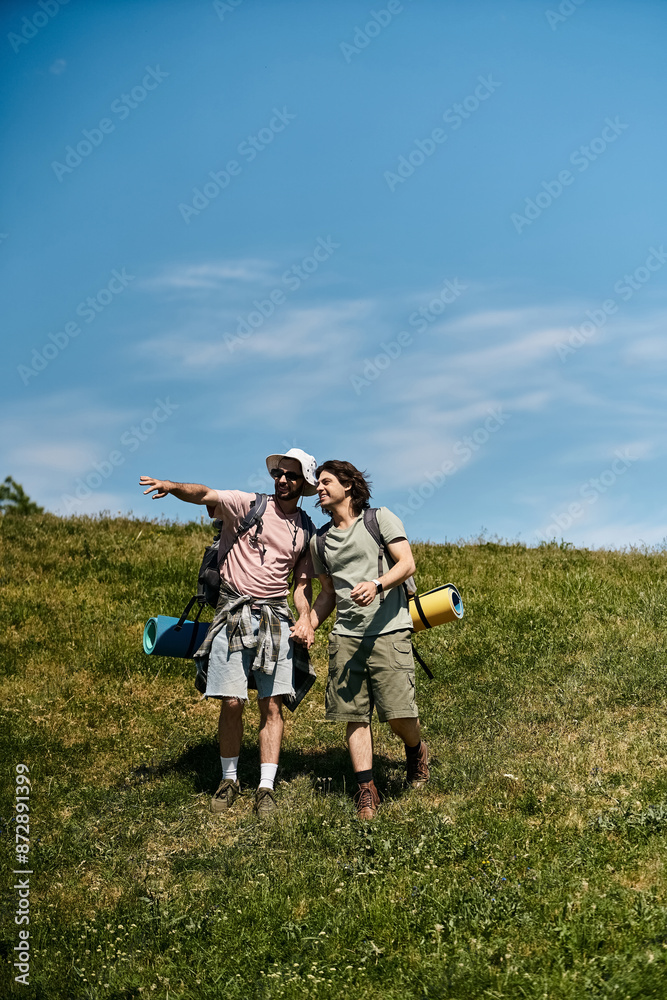 Fototapeta premium A young gay couple hikes through a grassy field under a sunny blue sky.