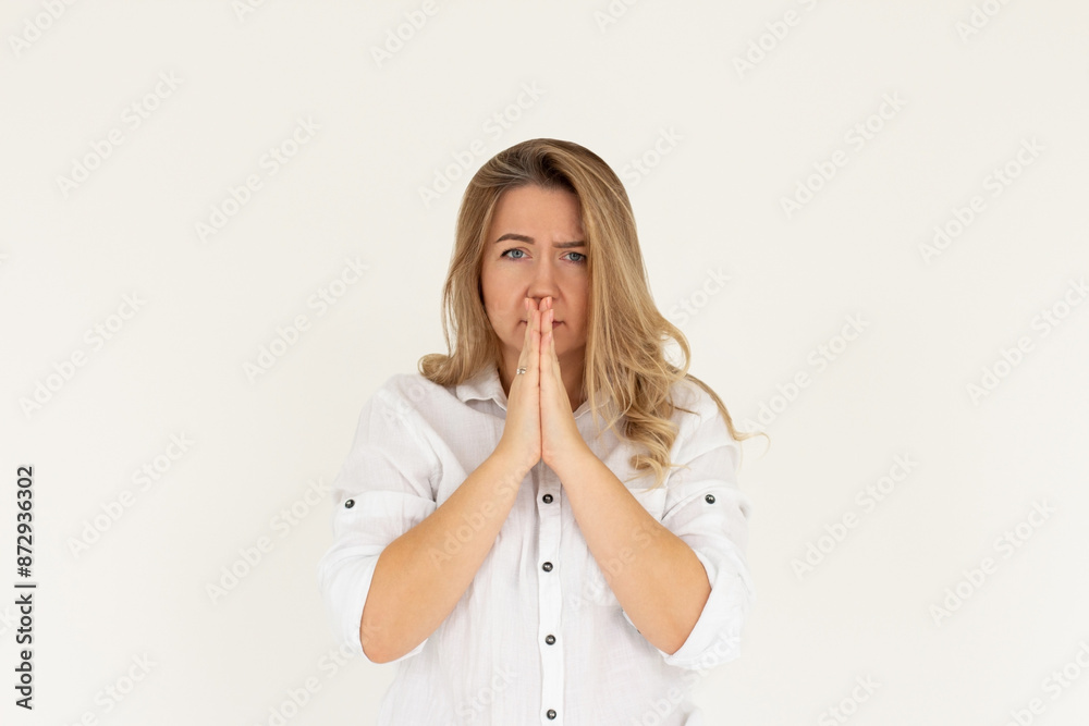 Portrait of imploring girl tilts head, purses lips, looks sadly at camera, makes praying gesture, asks for help and support, begs boyfriend for apologize, wears blue t-shirt, isolated on white wall Portrait of imploring girl tilts head, purses lips, looks sadly at camera, makes praying gesture, asks for help and support, begs boyfriend for apologize, wears blue t-shirt, isolated on white wall