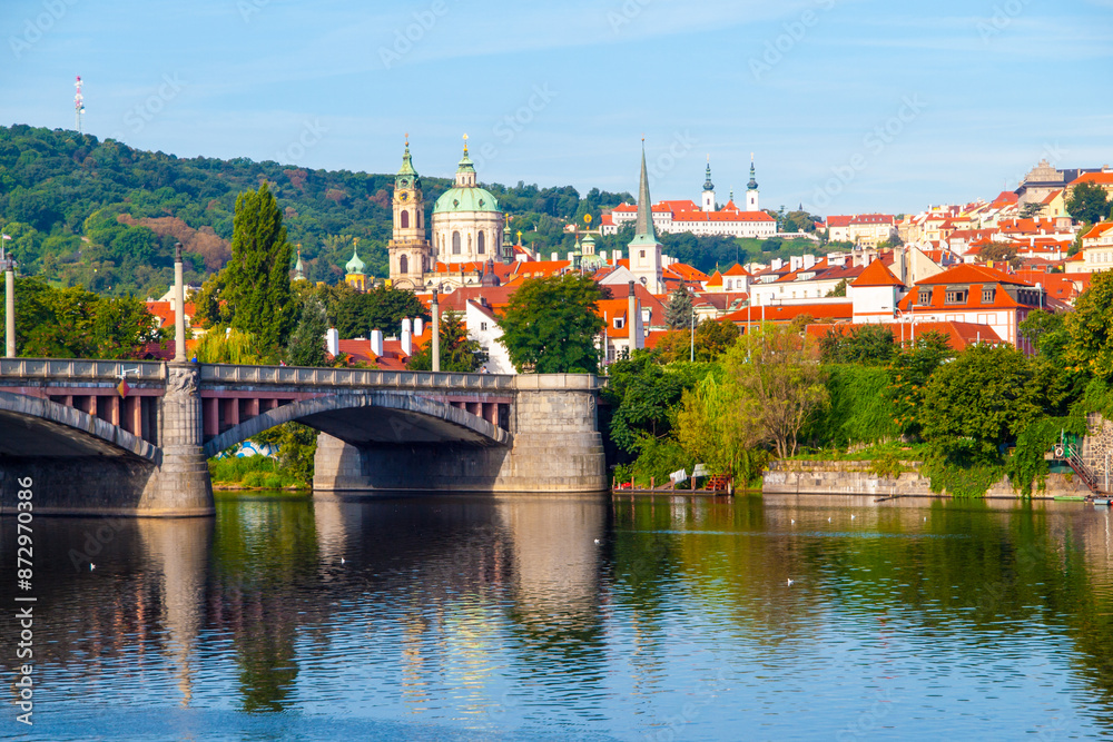 Fototapeta premium A stone bridge spans the Vltava River in Prague, Czechia. Red-roofed buildings line the hills in the background, with a church spire visible.
