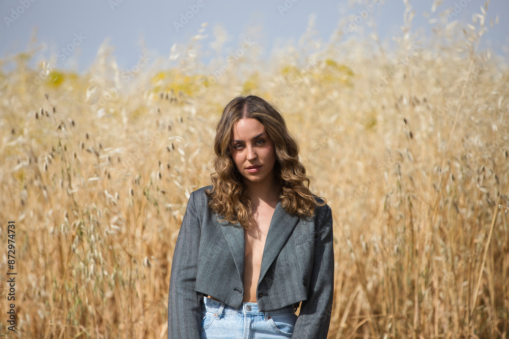 pretty young blonde woman with light eyes is dressed in grey jacket, jeans, boots sitting on a stool in a field planted with oats. The woman makes different expressions. pretty young blonde woman with light eyes is dressed in grey jacket, jeans, boots sitting on a stool in a field planted with oats. The woman makes different expressions.