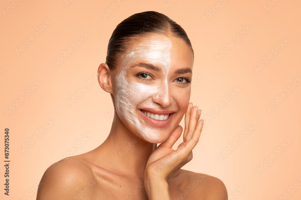 Close-up studio shot of happy European woman applying face mask on half of her face, posing against beige background, smiling at camera Close-up studio shot of happy European woman applying face mask on half of her face, posing against beige background, smiling at camera