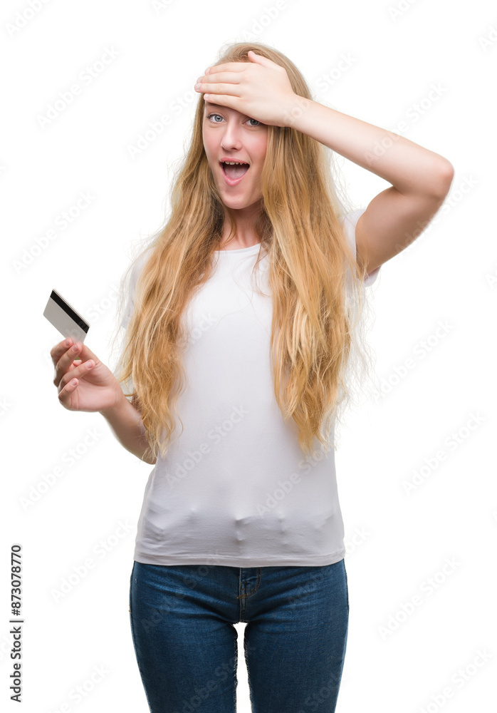 Naklejka premium Blonde teenager woman holding credit card stressed with hand on head, shocked with shame and surprise face, angry and frustrated. Fear and upset for mistake.