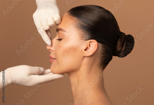 Plastic surgery and beauty concept. Surgeon touching woman nose, consulting before rhinoplasty operation on brown background, side view photo