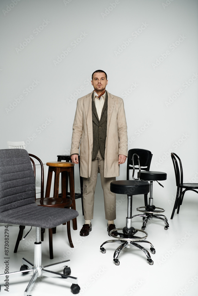 A stylish man in a beige coat stands confidently amidst a collection of chairs.