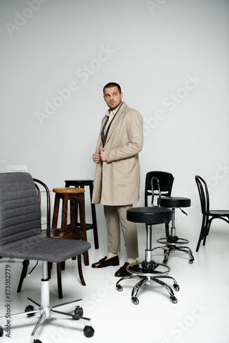 A confident man exudes timeless elegance in a room with varied chairs.