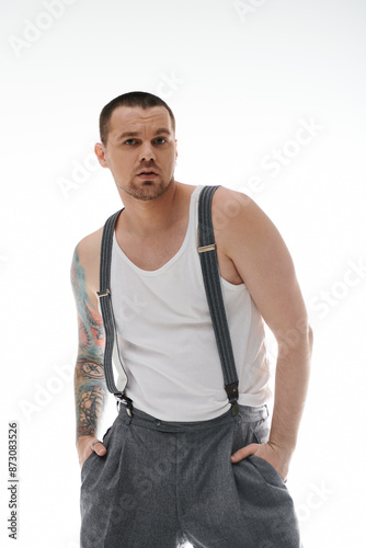 A man with tattoos on his arm poses in a white tank top and suspenders.
