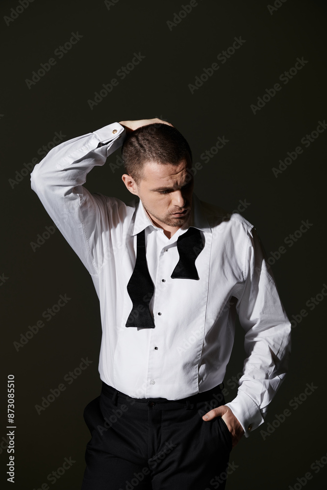 A man in a white shirt and black bowtie stands against a dark background, lost in thought. A man in a white shirt and black bowtie stands against a dark background, lost in thought.