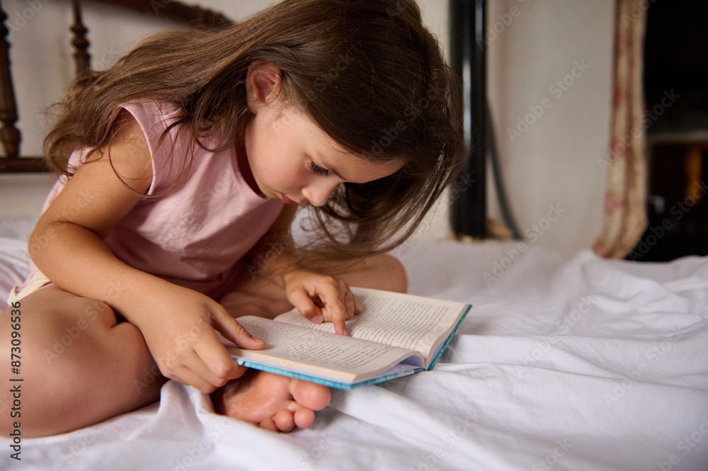 Close-up view of a cute child girl dressed in pink pajamas, running her finger along the line of sentences, reading book in her bedchamber. She looks concentrated, serious and focused Close-up view of a cute child girl dressed in pink pajamas, running her finger along the line of sentences, reading book in her bedchamber. She looks concentrated, serious and focused