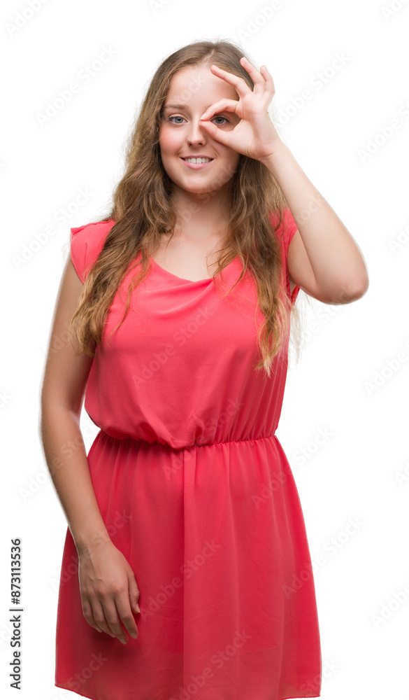 Young blonde woman wearing pink dress with happy face smiling doing ok sign with hand on eye looking through fingers Young blonde woman wearing pink dress with happy face smiling doing ok sign with hand on eye looking through fingers