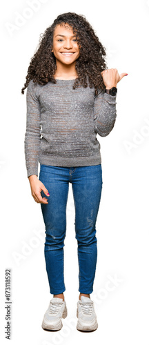 Young beautiful woman with curly hair wearing grey sweater smiling with happy face looking and pointing to the side with thumb up.