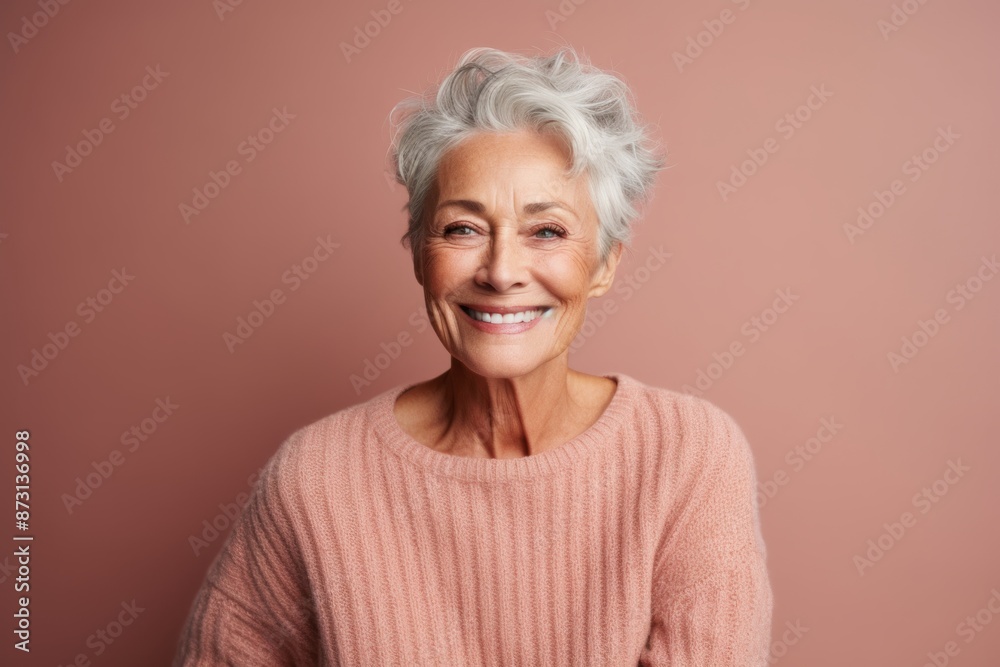 Portrait of a grinning woman in her 70s wearing a cozy sweater isolated in pastel brown background Portrait of a grinning woman in her 70s wearing a cozy sweater isolated in pastel brown background