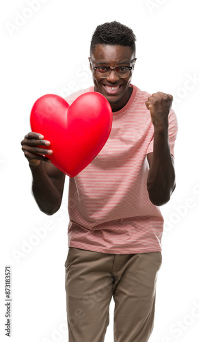 Young african american man holding red heart screaming proud and celebrating victory and success very excited, cheering emotion