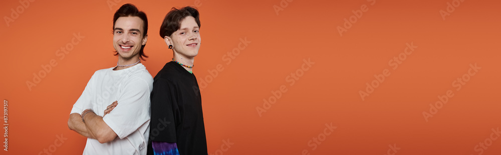Two young friends stand back-to-back, smiling brightly against a vibrant orange backdrop. Two young friends stand back-to-back, smiling brightly against a vibrant orange backdrop.