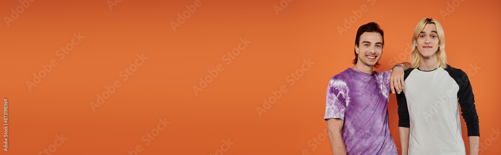 Fototapeta premium Two young queer friends in colorful clothing pose against an orange background.
