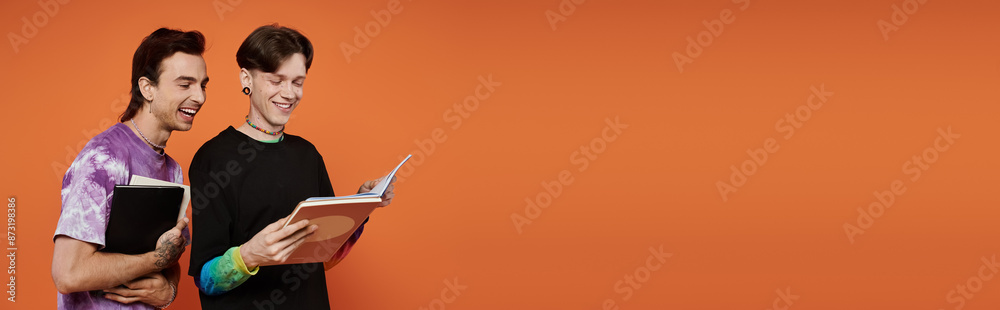 Two young queer friends in stylish attire smile while reading books against an orange background. Two young queer friends in stylish attire smile while reading books against an orange background.