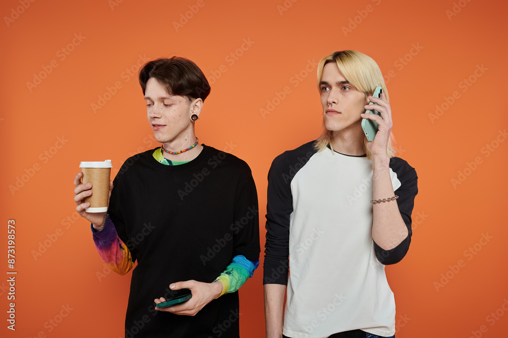 Two young queer friends pose in vibrant attire against an orange backdrop. Two young queer friends pose in vibrant attire against an orange backdrop.