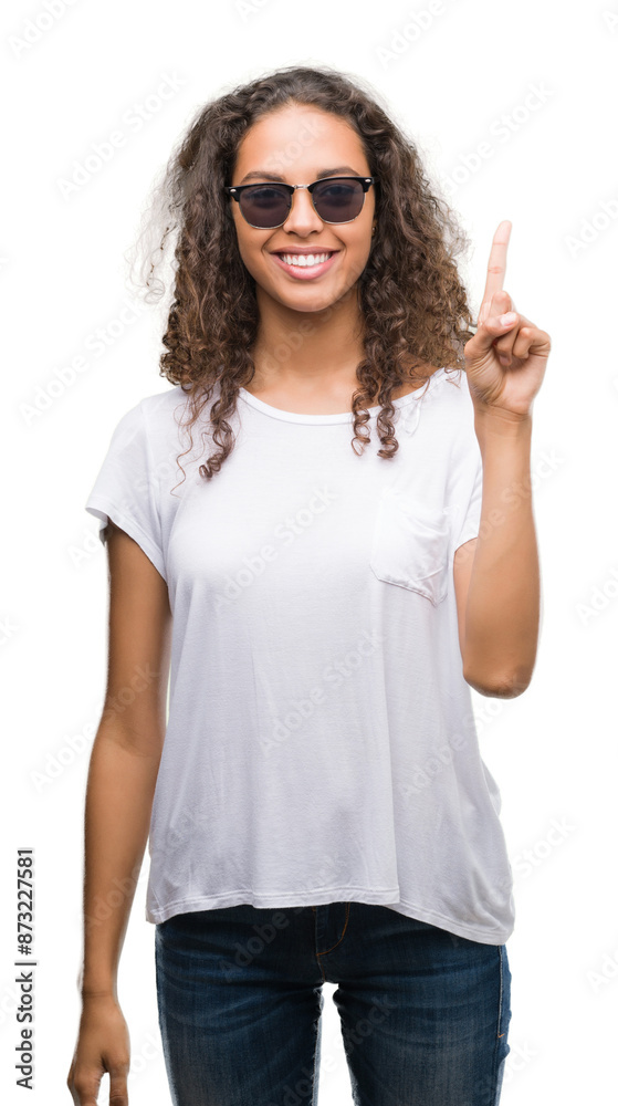 Young hispanic woman wearing sunglasses showing and pointing up with finger number one while smiling confident and happy.