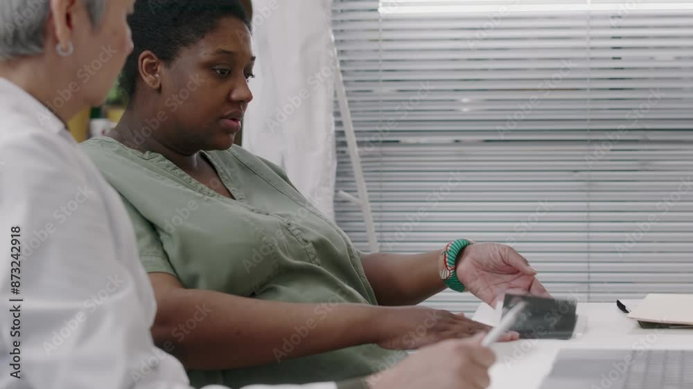 Medium tilt of young pregnant African American woman sitting at table with female unrecognizable gynecologist, typing on laptop while discussing birth plan and fetal ultrasound results Medium tilt of young pregnant African American woman sitting at table with female unrecognizable gynecologist, typing on laptop while discussing birth plan and fetal ultrasound results