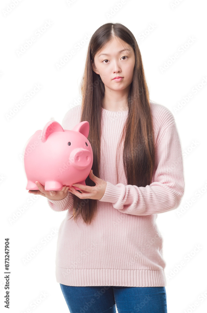 Young Chinese woman over isolated background holding piggy bank with a confident expression on smart face thinking serious Young Chinese woman over isolated background holding piggy bank with a confident expression on smart face thinking serious