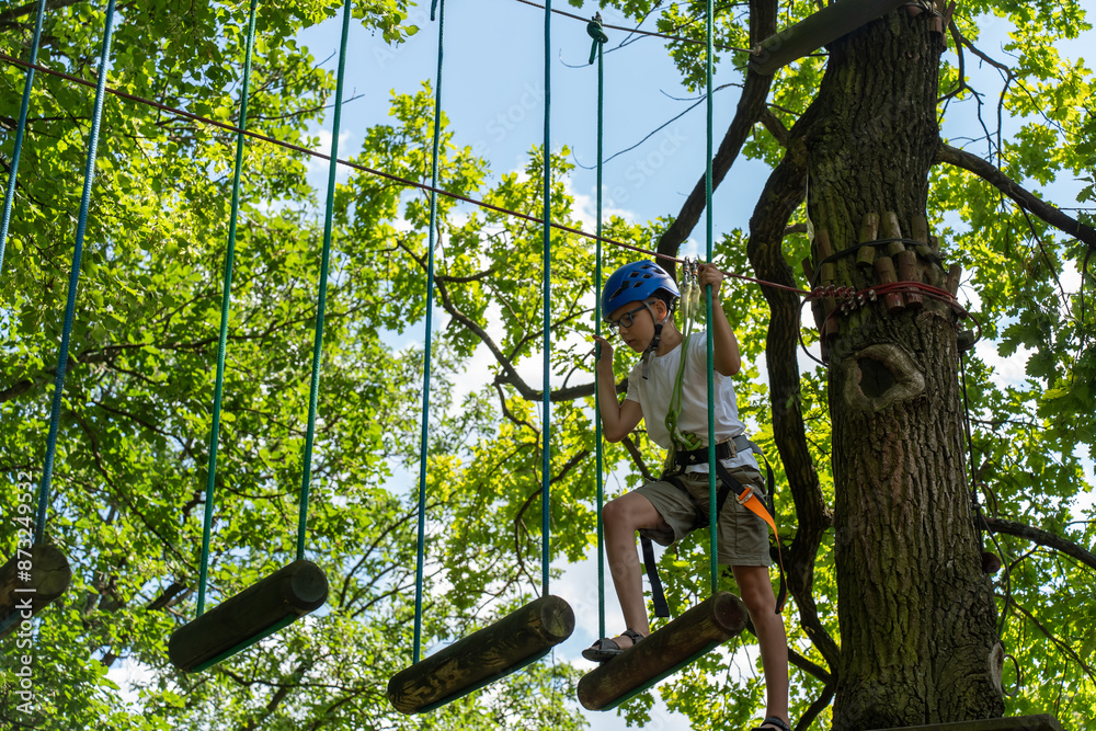 Obraz premium Boy exploring an outdoor playground with ropes and wooden obstacles. the child is wearing a helmet and safety gear, enjoying an adventure in nature on a sunny day. Rope trail in an adventure rope park