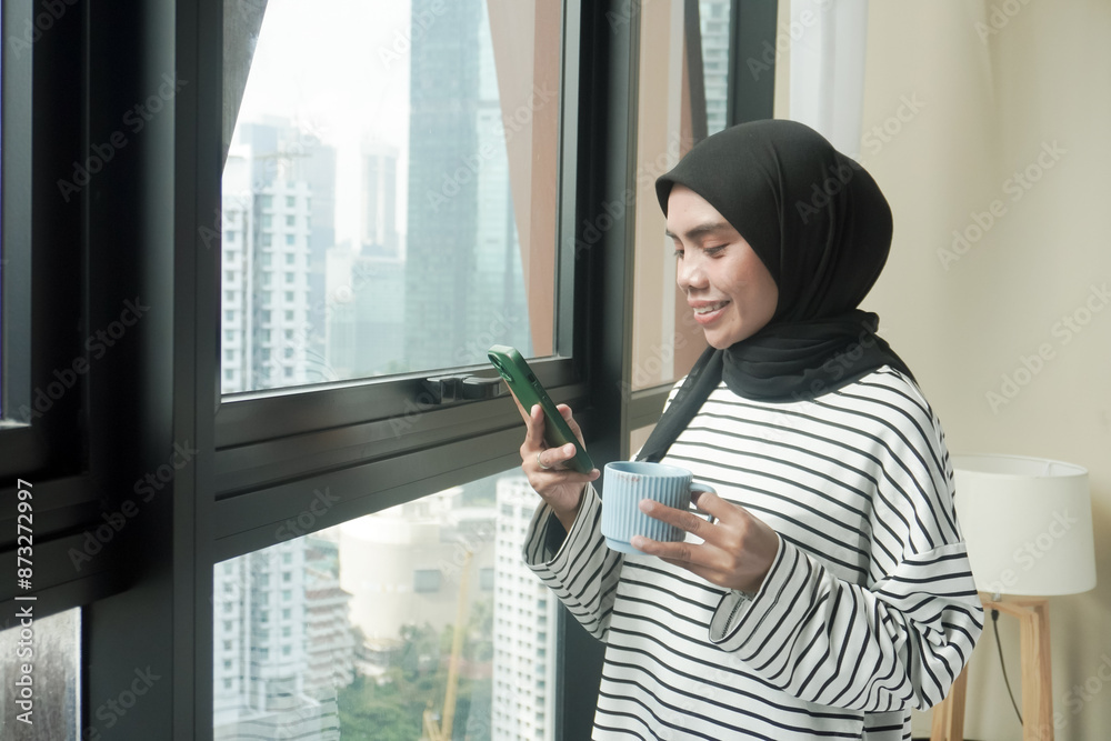 Fototapeta premium A woman is smiling and holding a cup of coffee while holding a cell phone. Concept of relaxation and enjoyment, as the woman is taking a break from her daily routine to enjoy a warm beverage