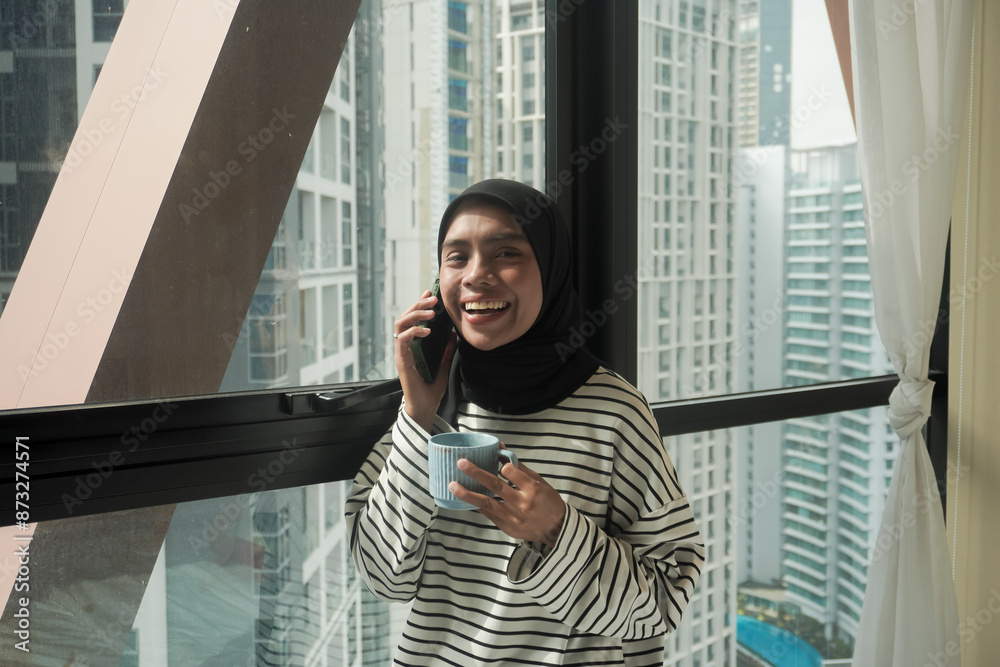 Fototapeta premium A woman is smiling and holding a cup of coffee while holding a cell phone. Concept of relaxation and enjoyment, as the woman is taking a break from her daily routine to enjoy a warm beverage