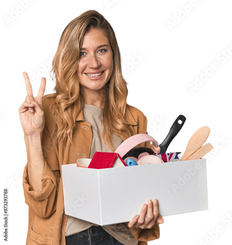 Young Caucasian blonde moving with a box joyful and carefree showing a peace symbol with fingers. photo